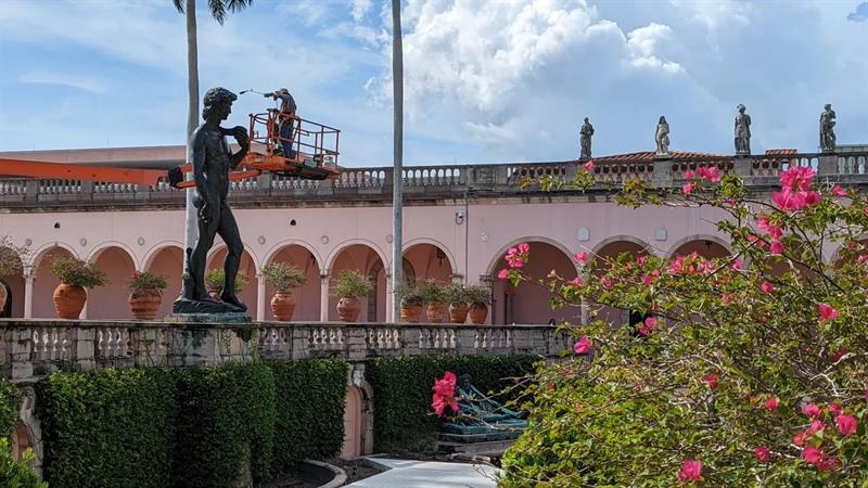A conservator stands on an elevated platform and uses a hose to rinse a statue in the courtyard of a museum.