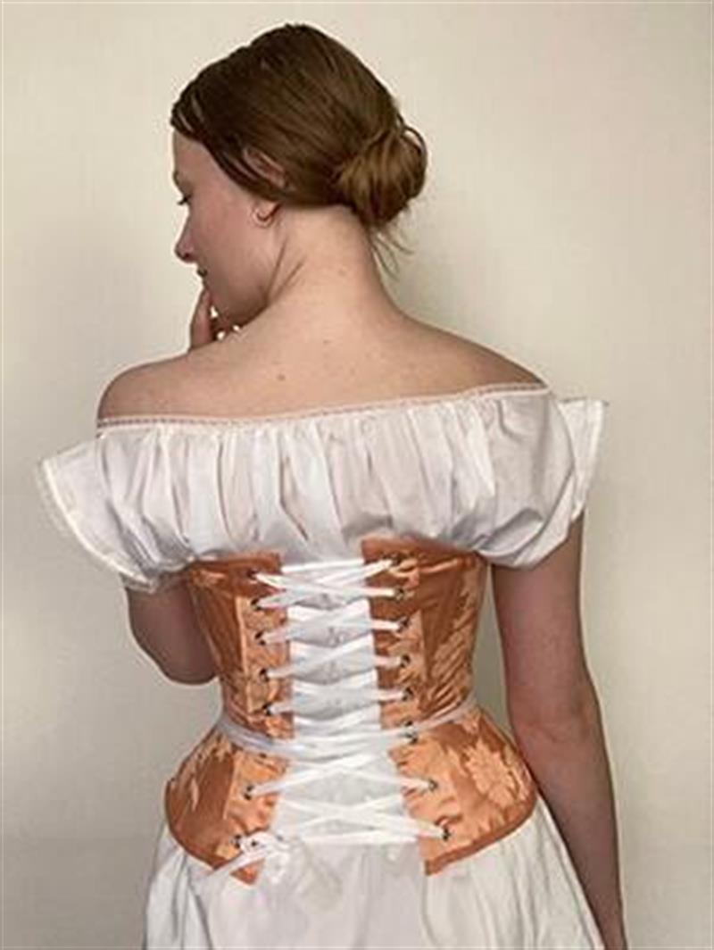 A student models historical garment reconstructions: a white dress with a peach-colored corset.