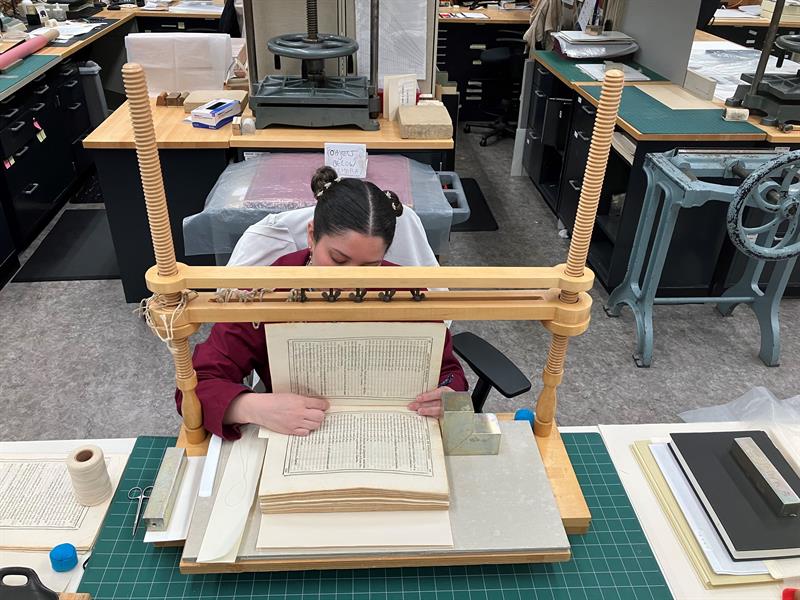 A student sits at a table and positions the book in a wooden stand used for book spine and binding construction.