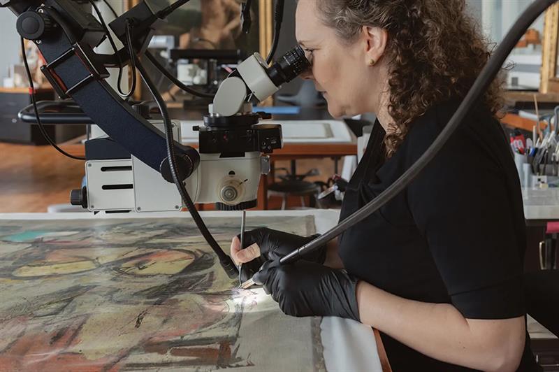 A conservator applies adhesive to loose paint, using a microscope and a small brush.
