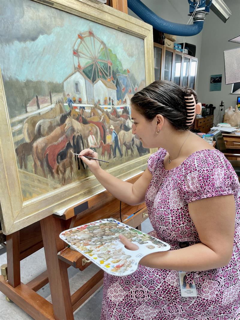 A student in a purple print dress sits at an easel and uses a small brush to inpaint an area of loss on a painting.