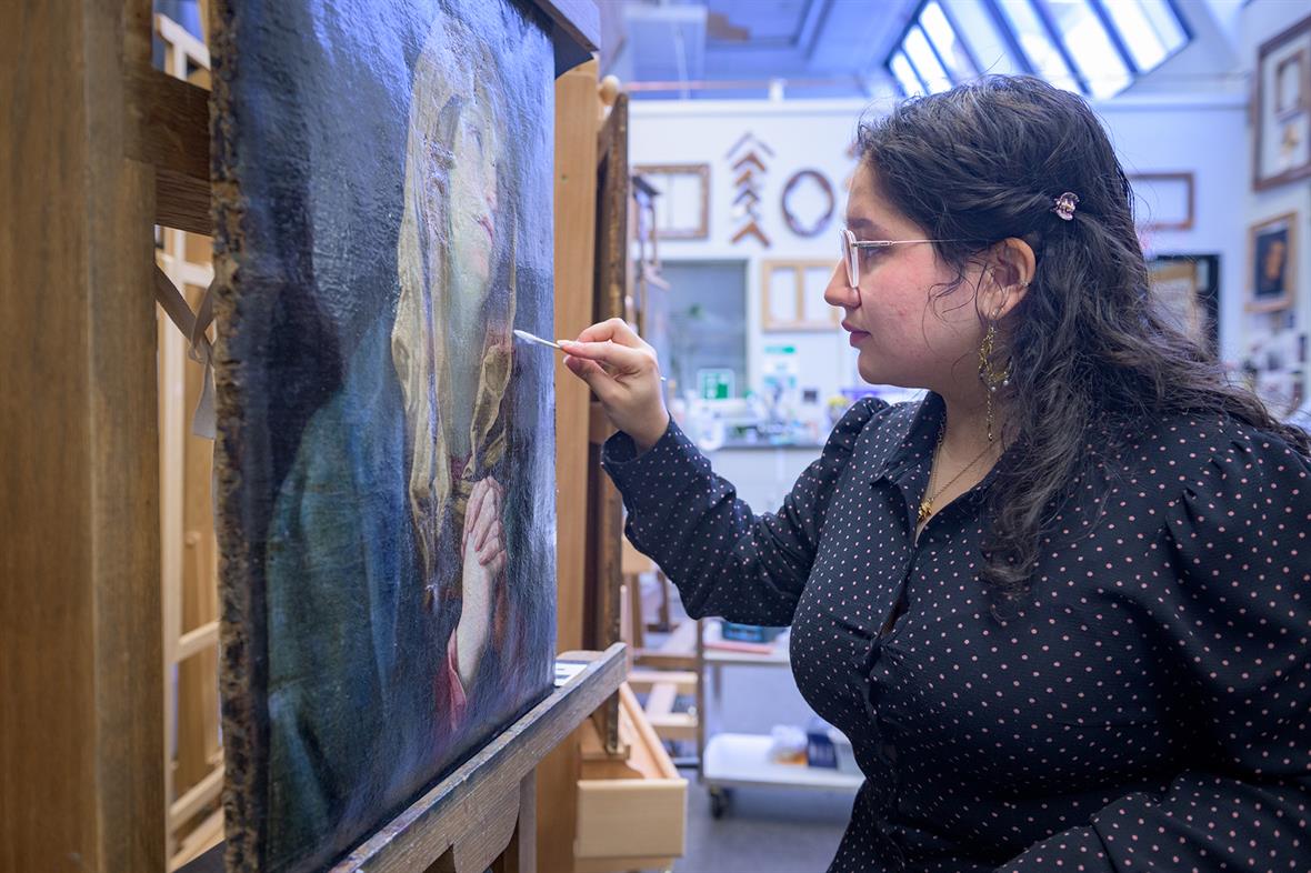 A student stands at an easel and touches a small cotton swab to the surface of a painting.