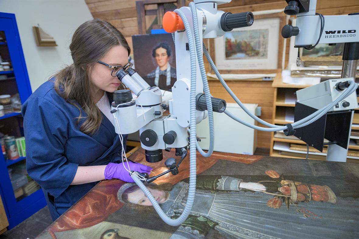 A student uses a microscope to look at a painting laying on  a table.