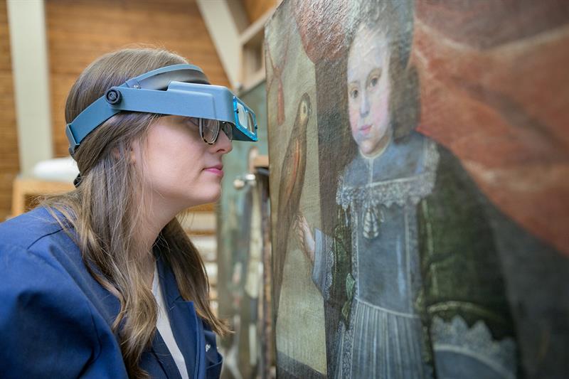 A student, wearing a magnifying visor, examines the surface of a painting set on an easel.