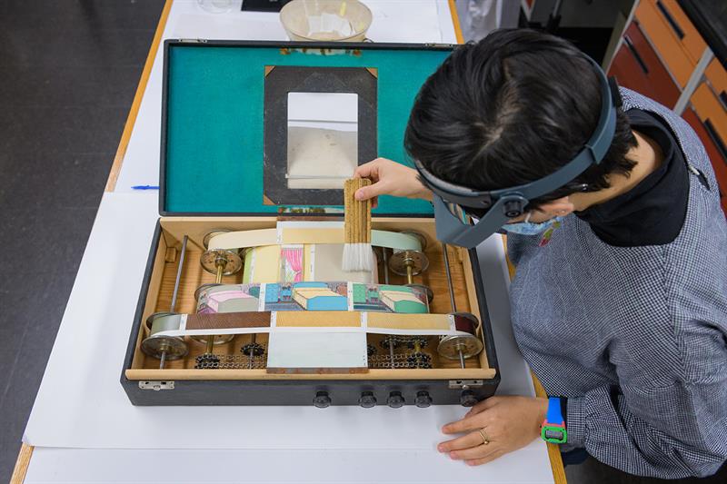 A student leans over the open Colorguide, which is sitting open on a table, and gently removes dust with a soft brush.