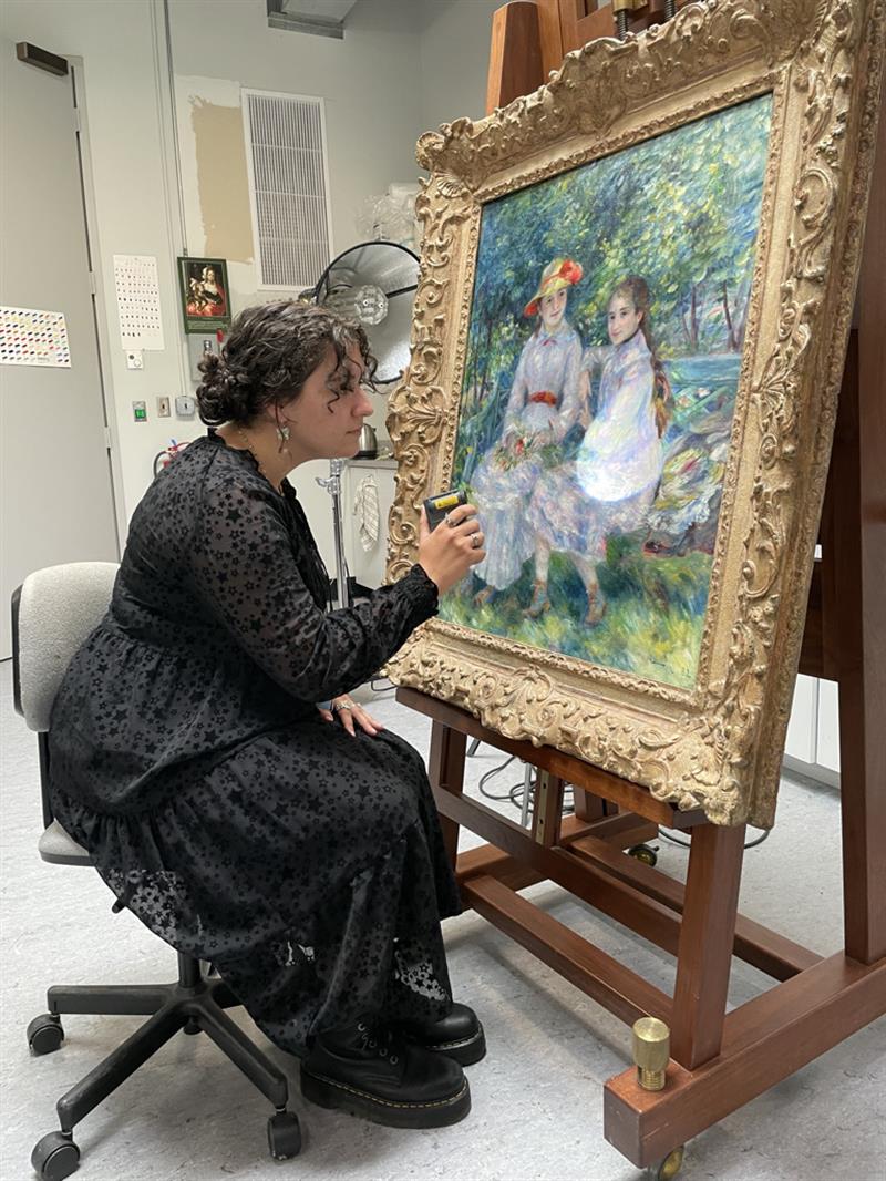A student in a long black dress sits in a chair and looks at a painting of two women.