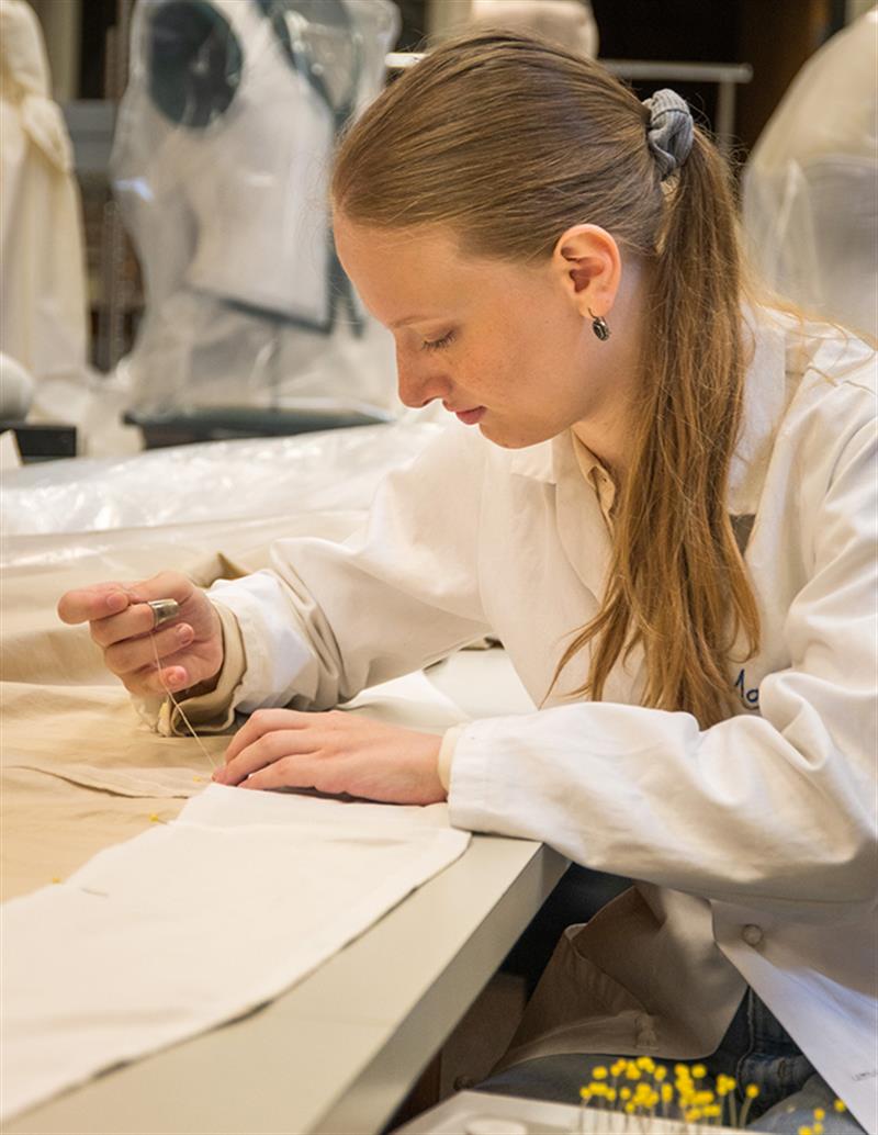 A student works at a table, stitching a seam in a muslin fabric.