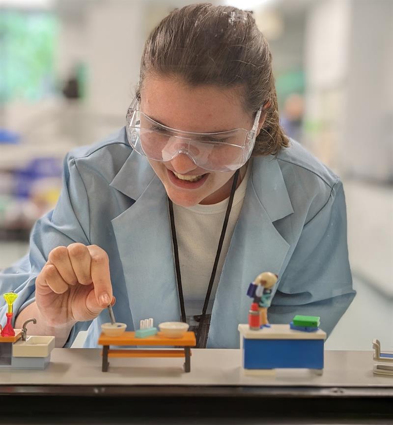 A student, wearing a lab coat and goggles, smiles as she plays with lab-themed toys.