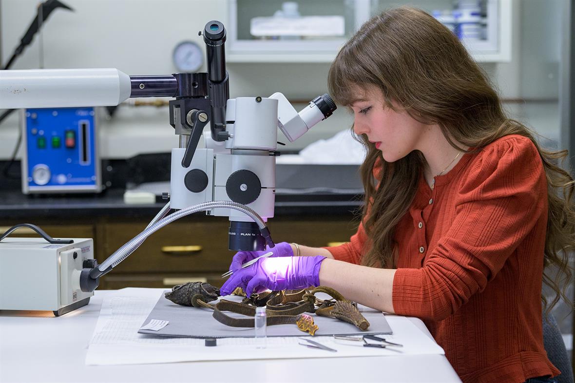 A student sits at a table and uses a microscope and small blade to take samples from the belt.