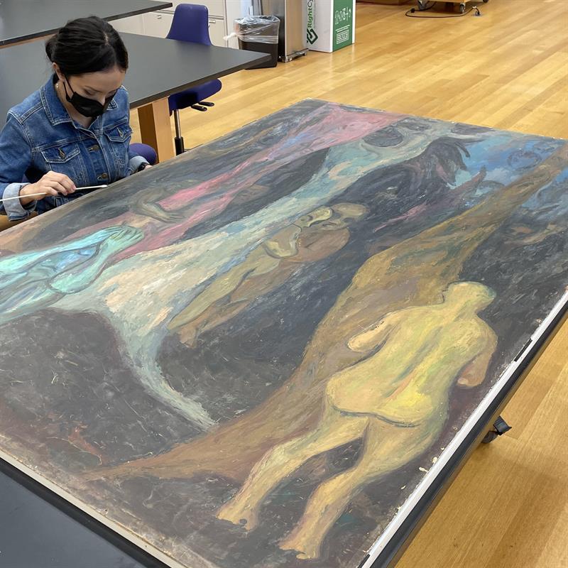 A student, wearing a mask, gently rolls a cotton swab over the edge of a large painting that is laying flat on a table.