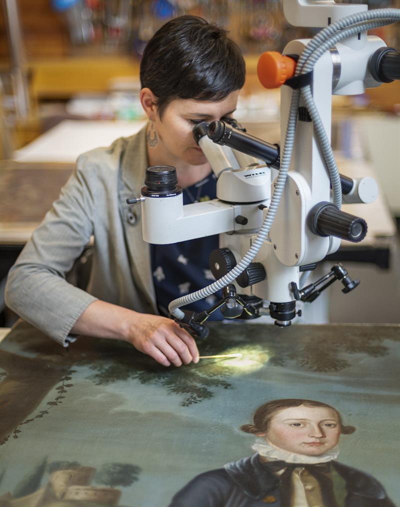 A student uses a microscope to look at a painting laying on a table.