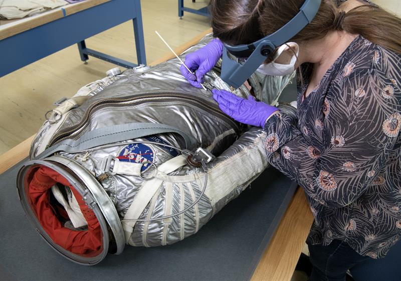 A conservator works on a spacesuit on a table, using a cotton swab to clean metal corrosion.