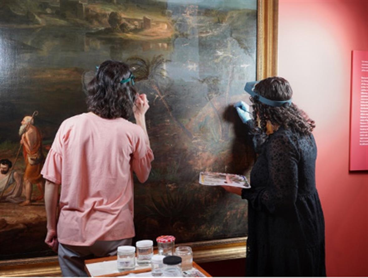 Two students stand in front of a painting in a gallery, and use small brush to repair problems with the varnish layer.