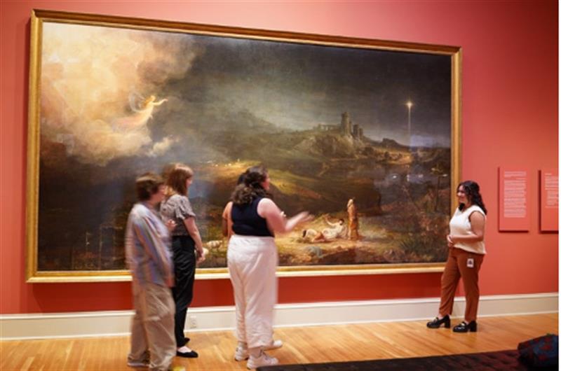 A group of students stand in a gallery, in front of a large painting.