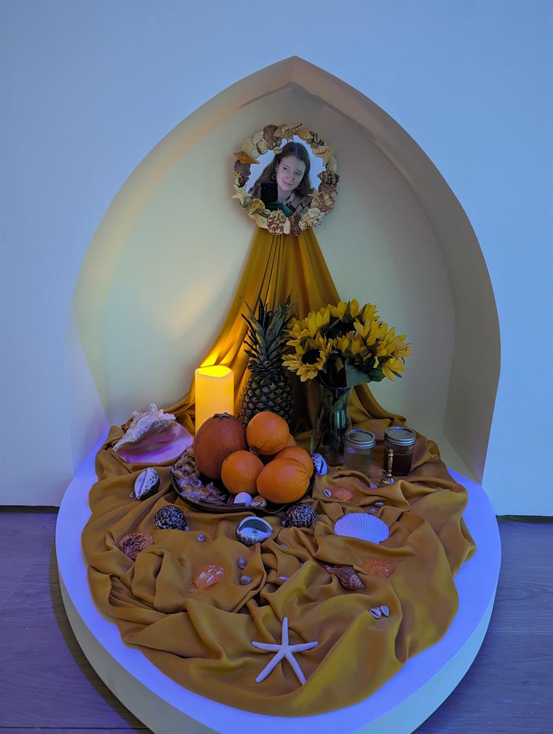 A mixed media artwork includes a figure, an eletric candle, fabrics, fruit, and shells.