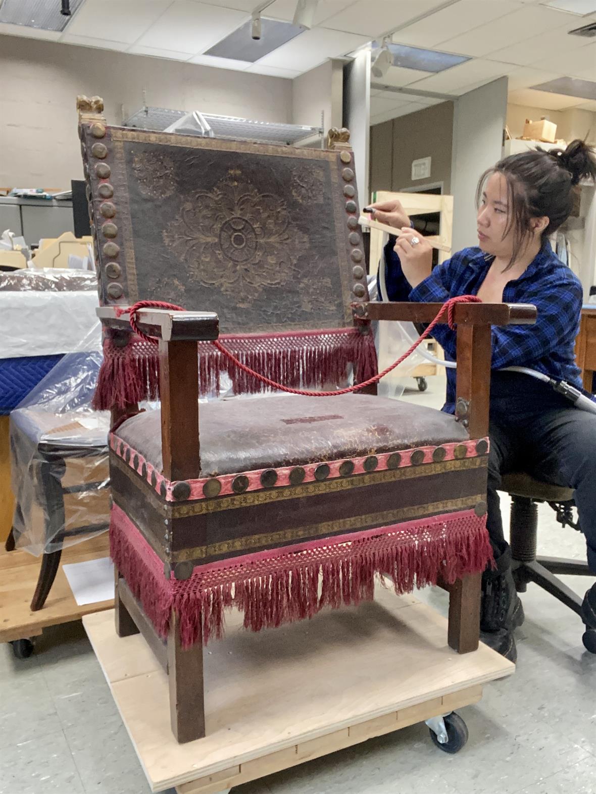 A student uses a soft brush and miniature vacuum to remove dust from the surface of an ornate chair.