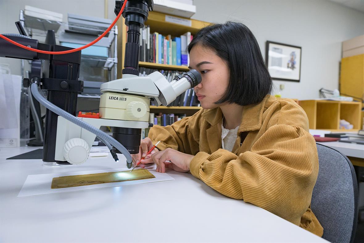 A student sits at a table and looks through a microscope to guide the placement of a tiny brush on the cover of the gazette.