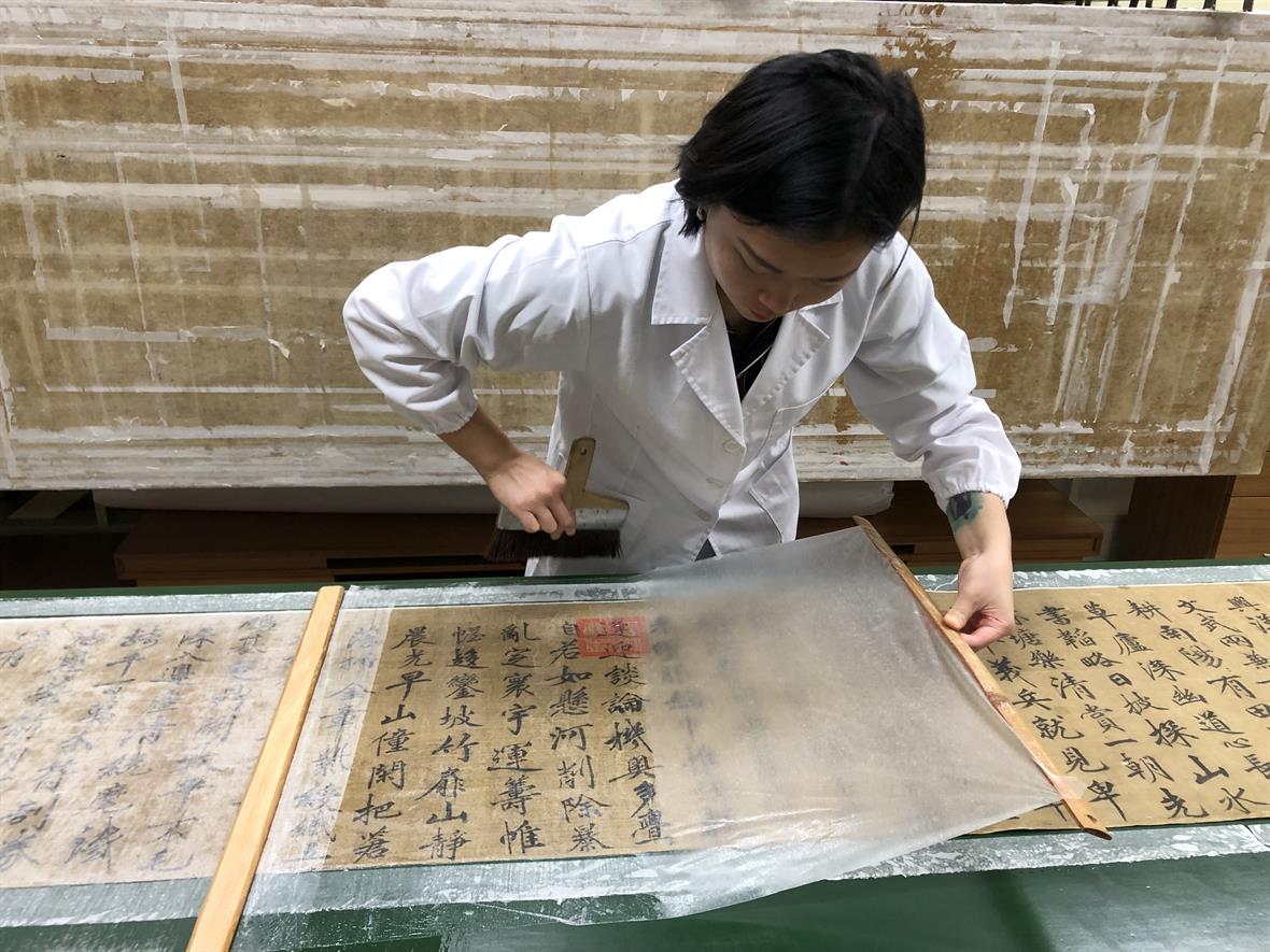 A student places thin tissue over the front of a delicate silk painting to support the fabric during treatment.