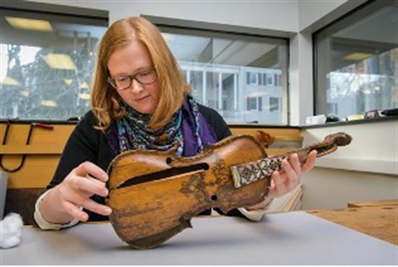 A student examines a wooden fiddle placed on a table.