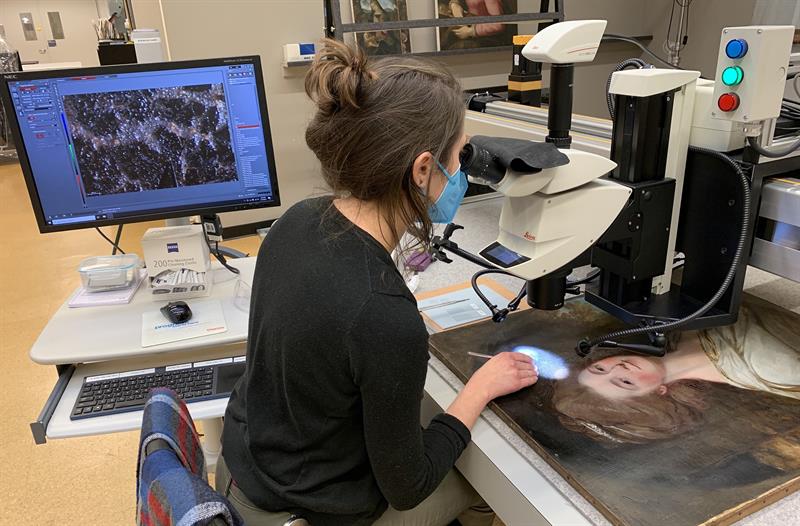 A student sits at a table and looks at a painting under a microscope. A nearby computer monitor shows the detail of the painting as seen through the microscope.