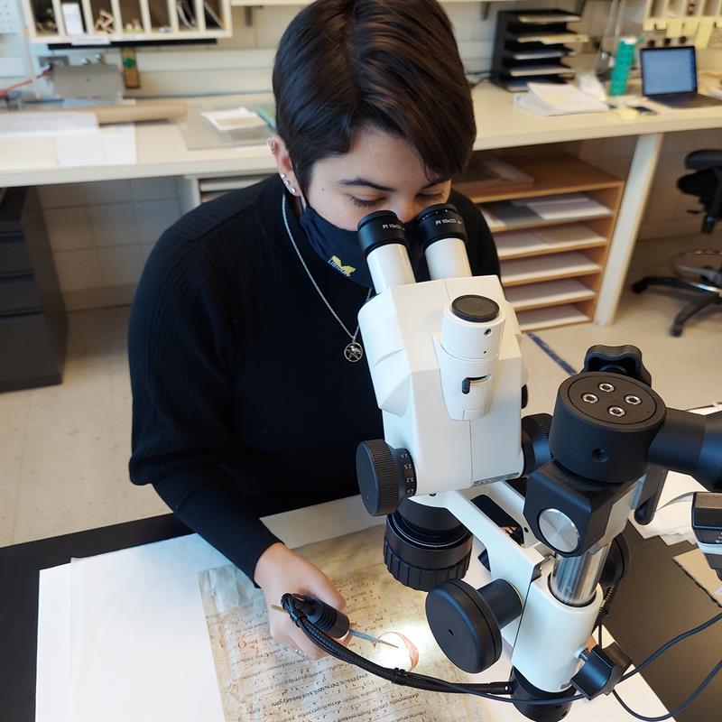 A student looks at a manuscript page using a microscope, while using a small brush to test for loose materials on the manuscript page.