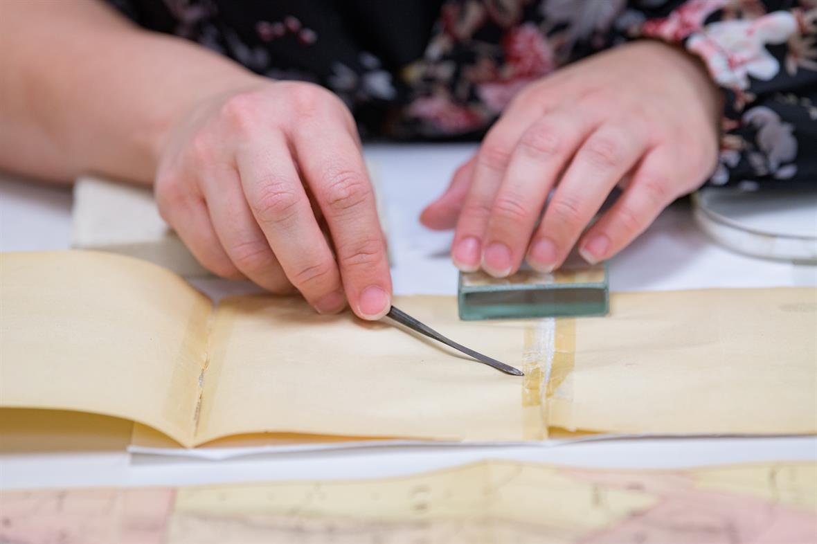 A student uses a glass weight to hold two pieces of the map in place while they use a small metal tool to lift the edge of old tape joining the papers.