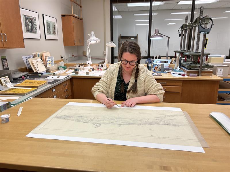 A woman wearing a sweater and glasses sits at a table and looks at a large map laid out on a piece of white blotter paper.