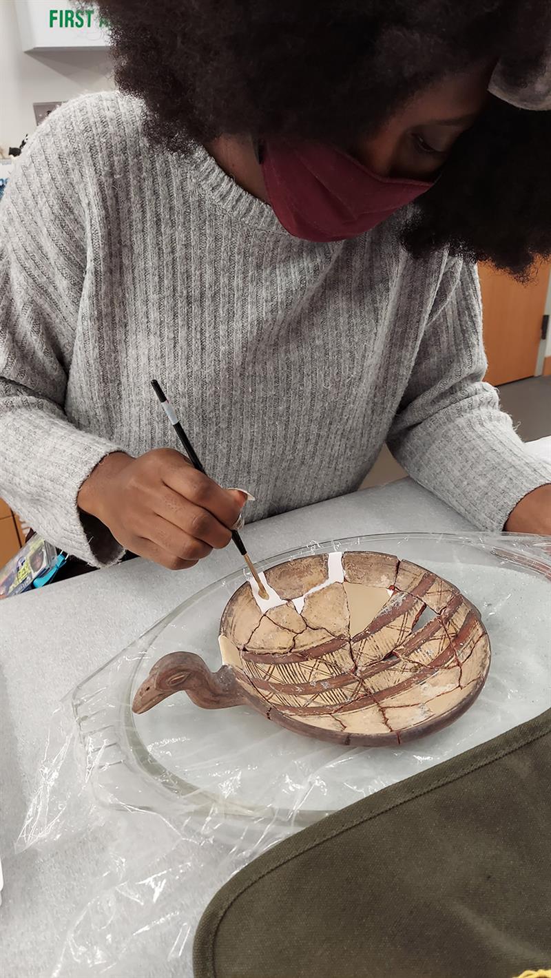 A student, wearing a mask, uses a small brush to tone white plaster that fills the cracks on a ceramic.