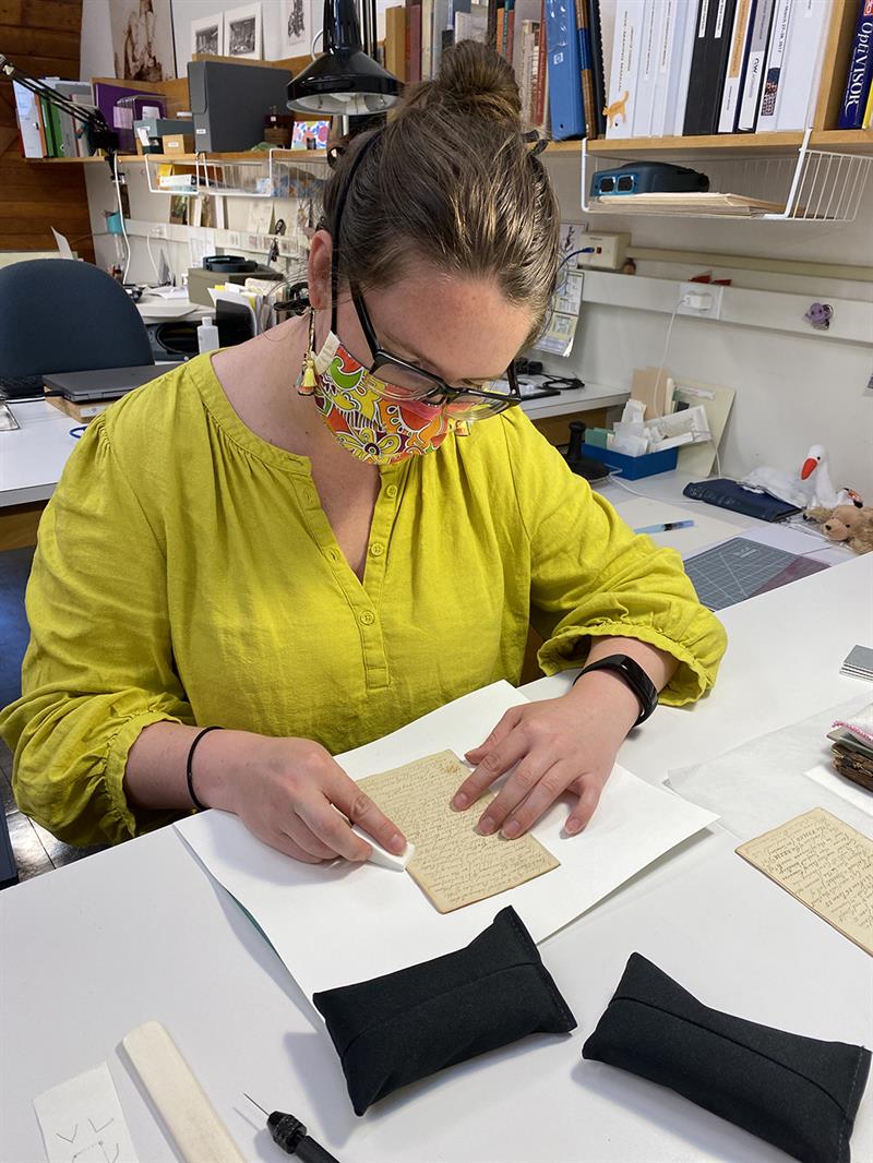 A student, wearing a mask, sits at a table and carefully touches a cosmetic sponge to the surface of a diary page.