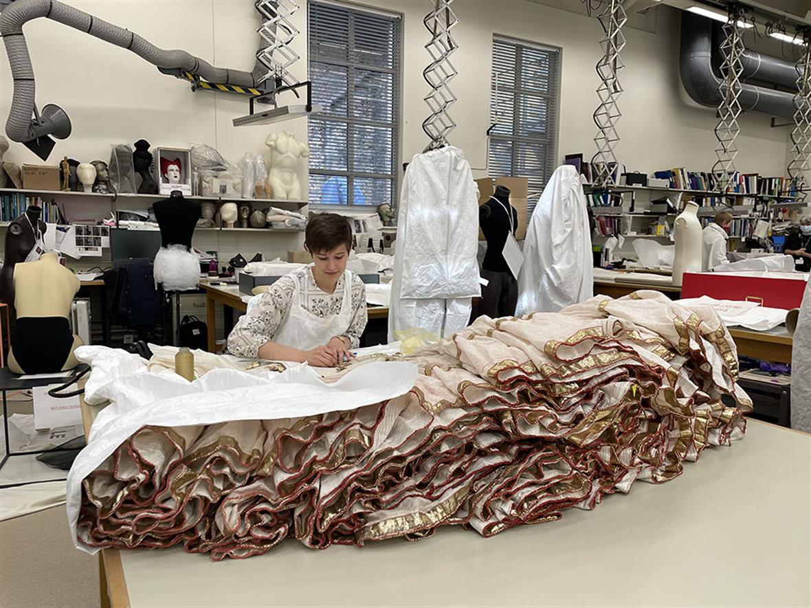 Student sits at a table in a conservation lab. The lab is full of mannequins; a multi-layered skirt lays on the table.