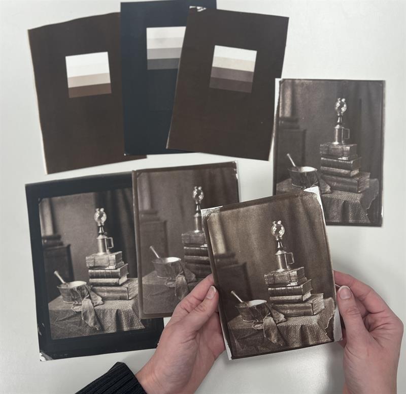 A student examines a photographic print, lifted above a selection of other prints on a table.
