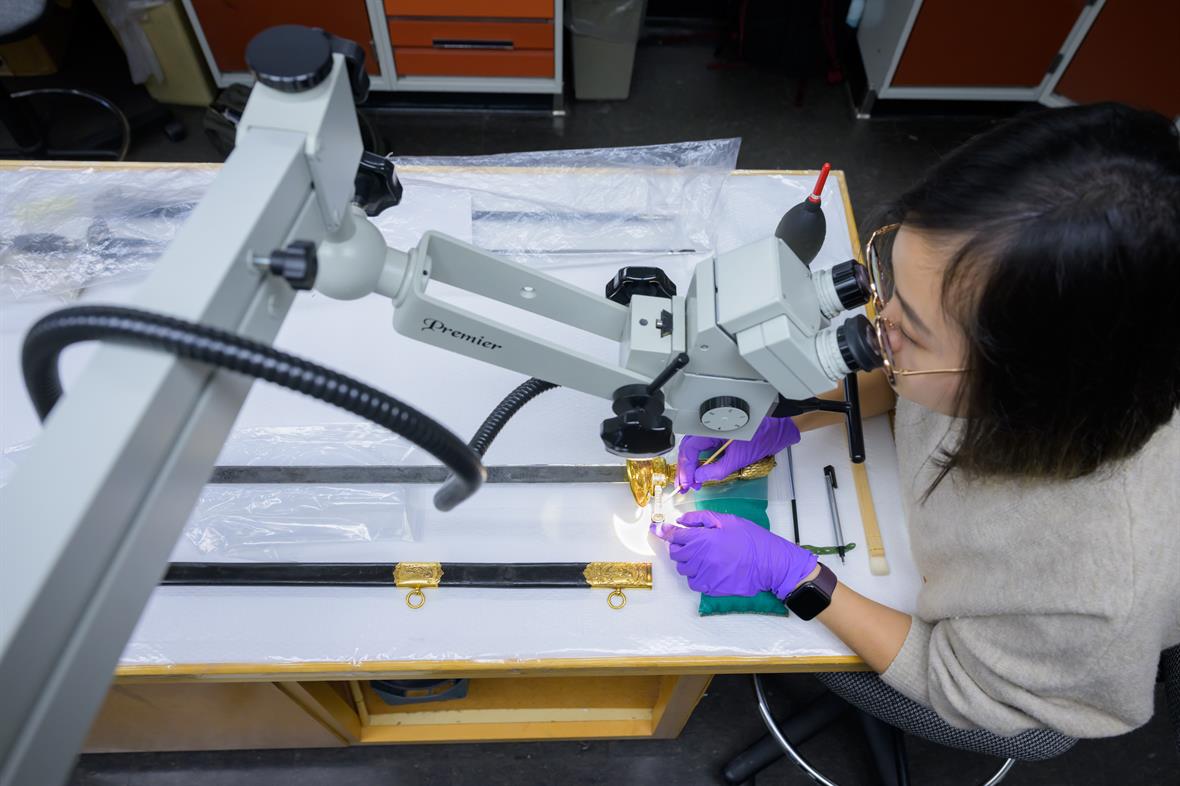 A student sits at a table and uses a microscope to guide tools across a sword hilt.
