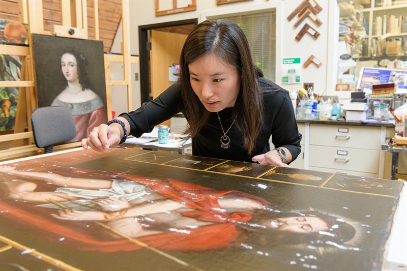 A student leans over a painting that is lying flat on a table. The student is using a microspatula to apply a white filling material to losses in the paint surface.