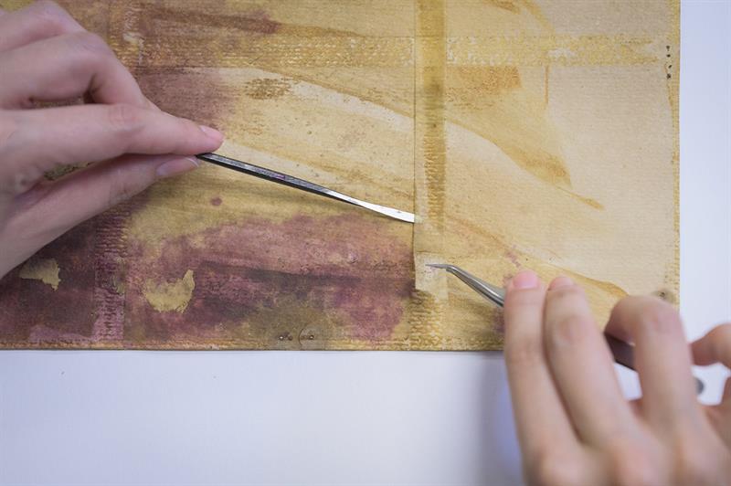 A close up of two hands using a microspatula and tweezers to lift the edge of an old, yellowed, brittle piece of tape from a piece of paper.