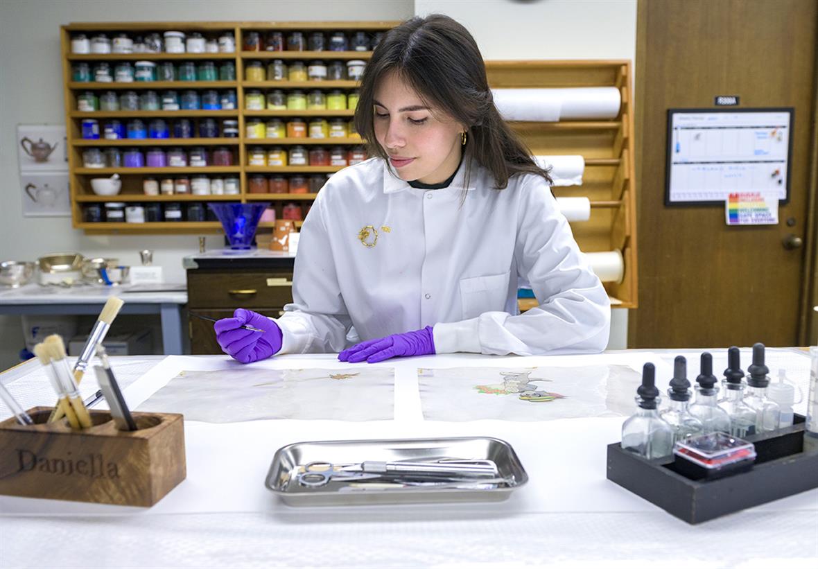 A student, wearing a lab coat and purple gloves, sits at a table. On the table are two animation cels, and trays of tools. Behind the student is a rack of colorful pigments in jars.