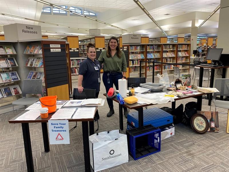 Two women stand in a library, behind two tables filled with collections care literature and materials.