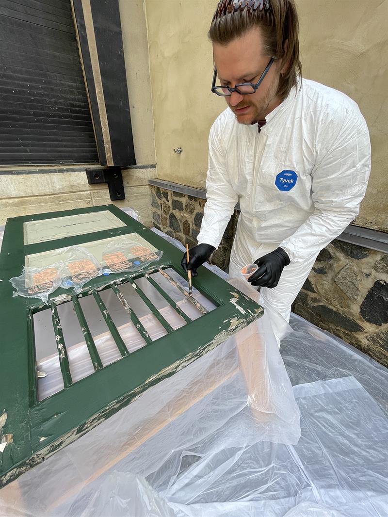 The student applying a thin putty to areas of paint loss on a green shutter.