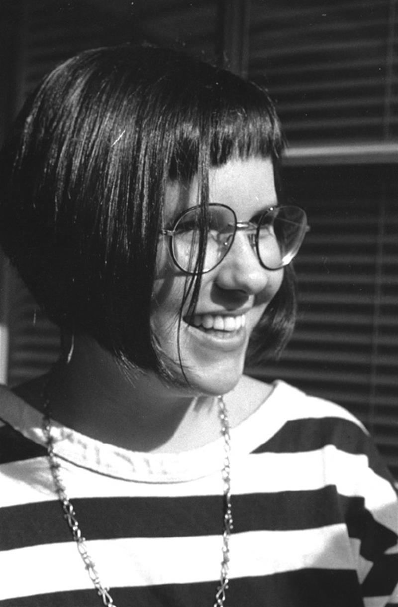 A woman smiles at the camera. She is wearing a striped shirt, glasses, and a necklace.