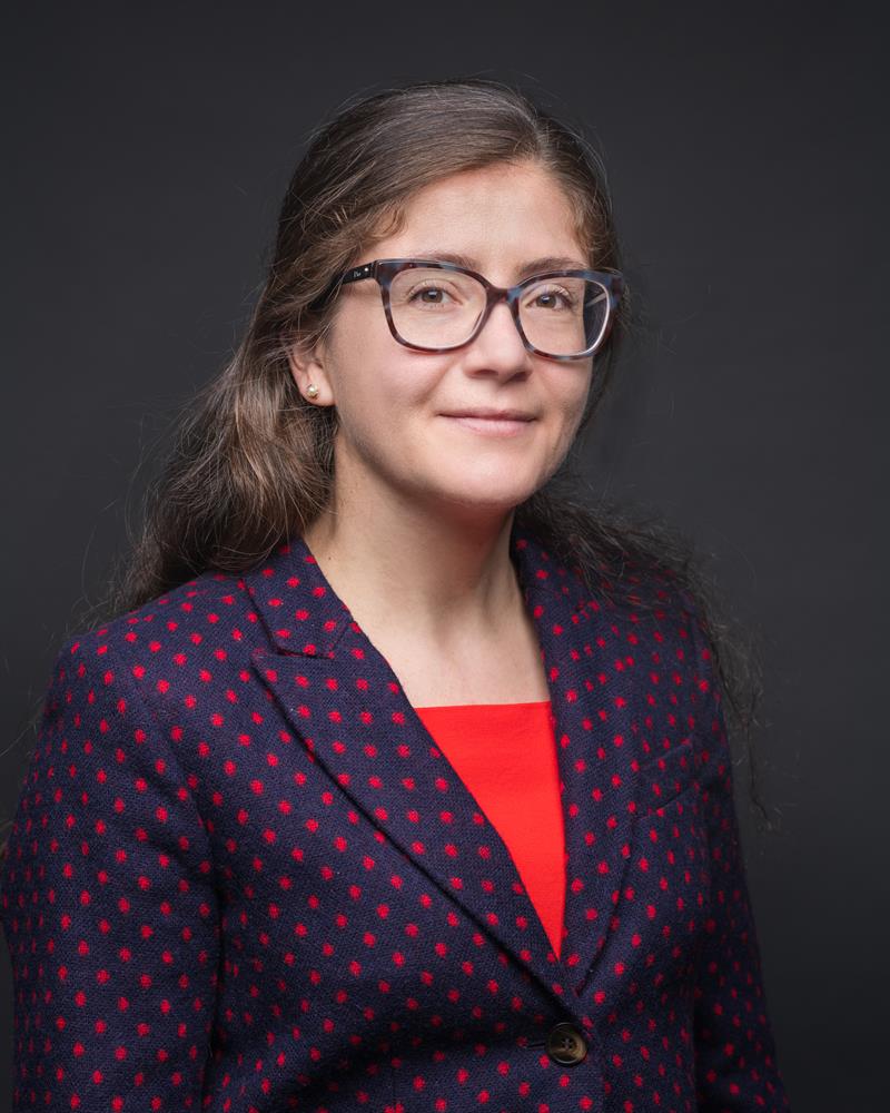 Portrait photo of a woman wearing glasses, a red shirt, and a blue suit jacket with red dots.