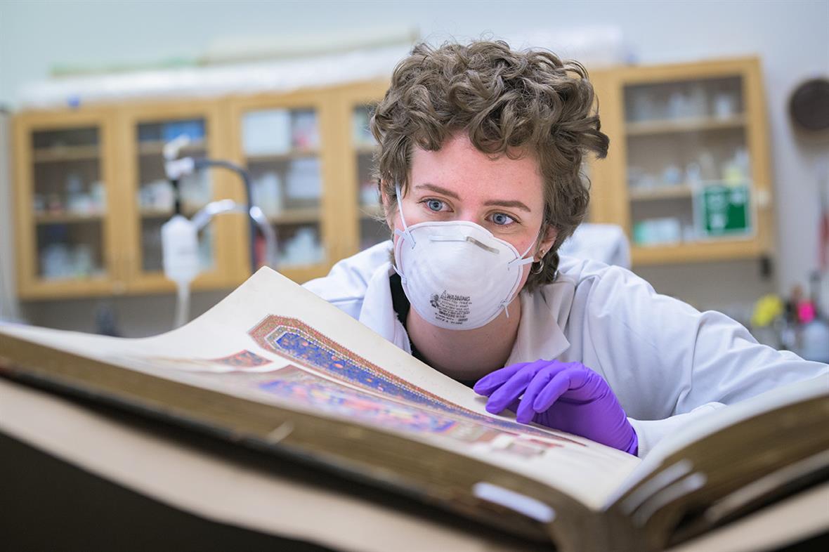 A student wearing a mask and gloves examines an oversized book open on a table.
