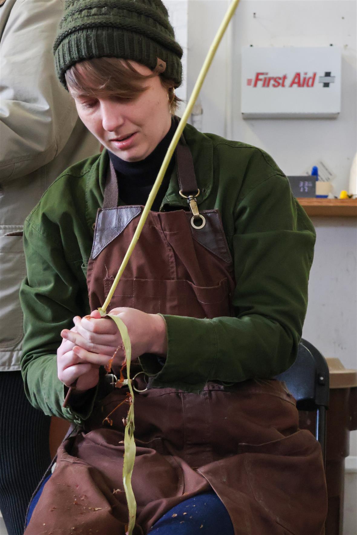 A student, wearing an apron, jacket, and hat, peels a piece of bark from a small branch.