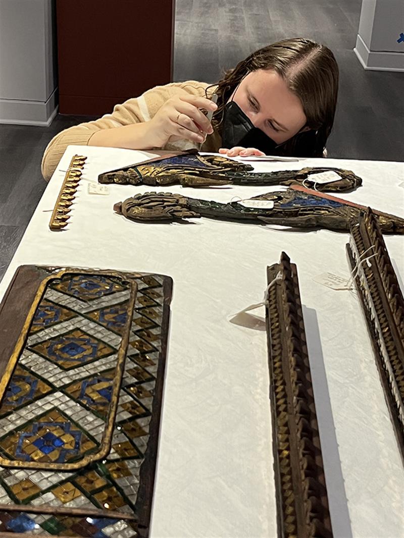 A student, wearing a mask, examines glass mosaics on a table.