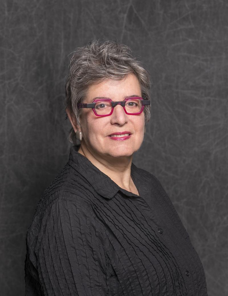 Portrait photograph of a women with short, grey hair. She is wearing a dark shirt and red-rimmed glasses.