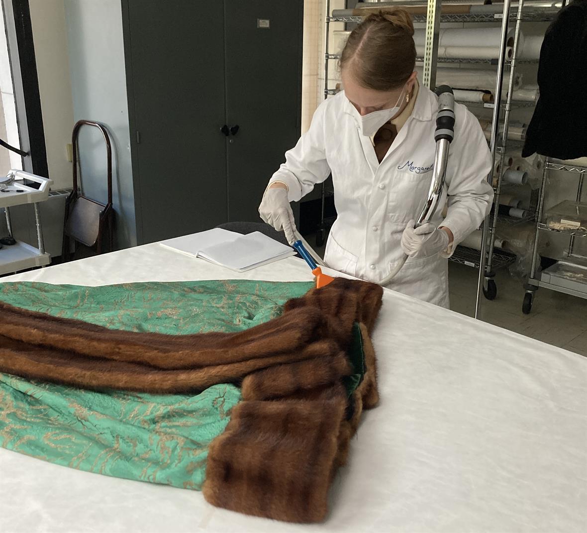 A student, wearing a lab coat and a mask, uses a hand-held vacuum to clean the fur trim on a textile.