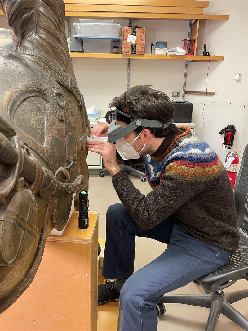 A student sit in front of a wooden horse, and uses magnifying glasses and a small scalpel to take a tiny piece of paint from the scupture.