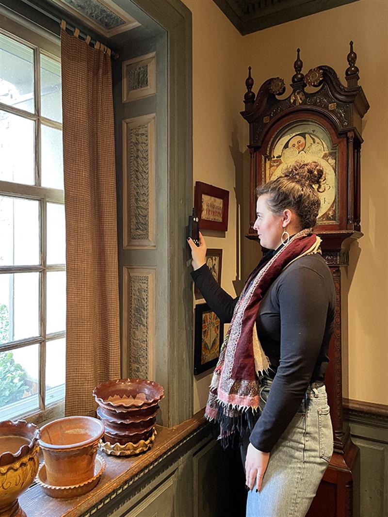 A student holds a light meter near some artwork by a window.