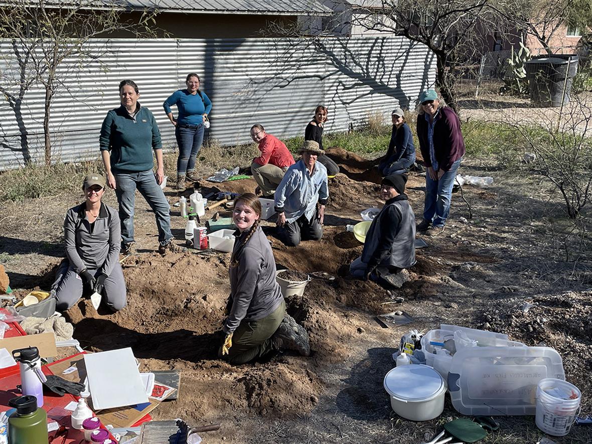 A group of students scattered around excavated areas of dirt, surrounded by plastic tubs and other tools for the excavation exercise.