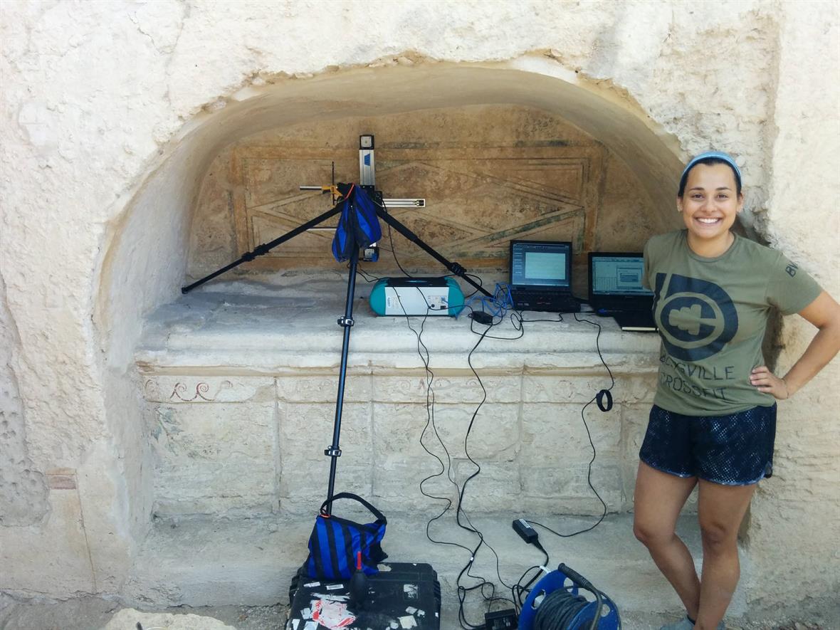  A woman in shorts and a t-shirt stands next to a wall painting inside a Grecian tomb. Analytical equipment and computers are set up along the edge of the painting.