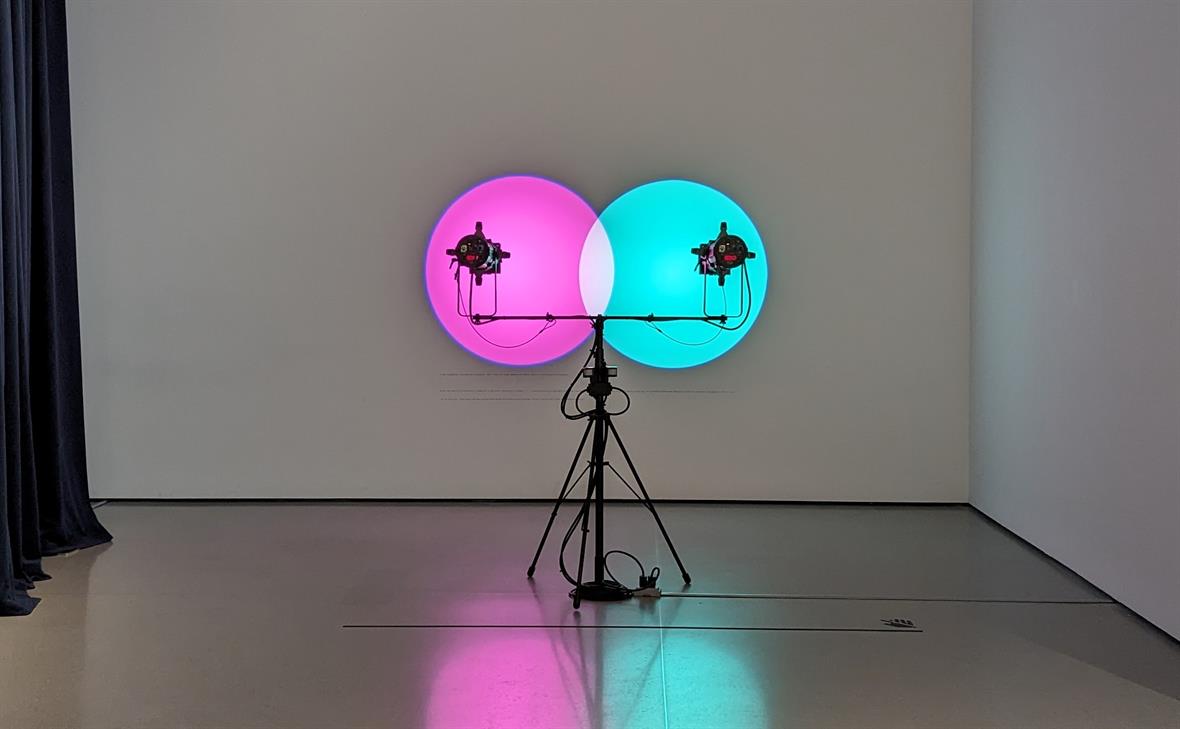 A tripod stands in a dimily lit room. Lights on either end of a horizontal pole on the tripod project pink and teal circles on a wall.