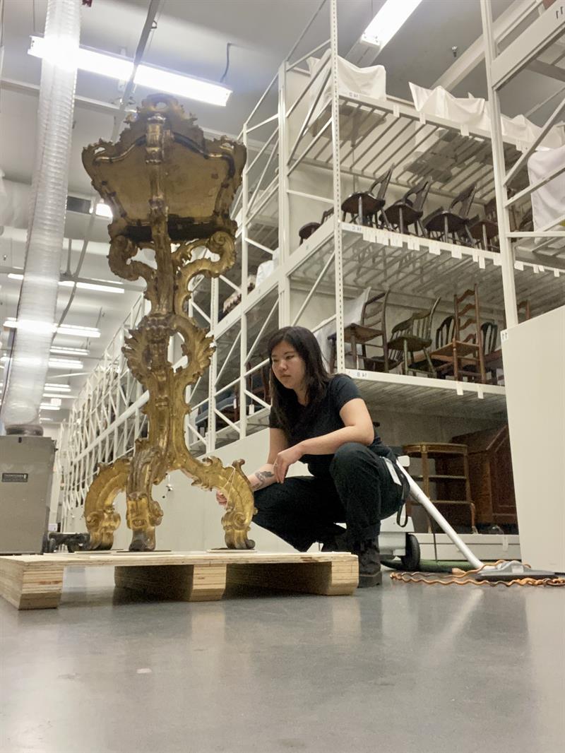 A student kneels to examine a gilded object on a platform. Rows of chairs fill shelves in the background.